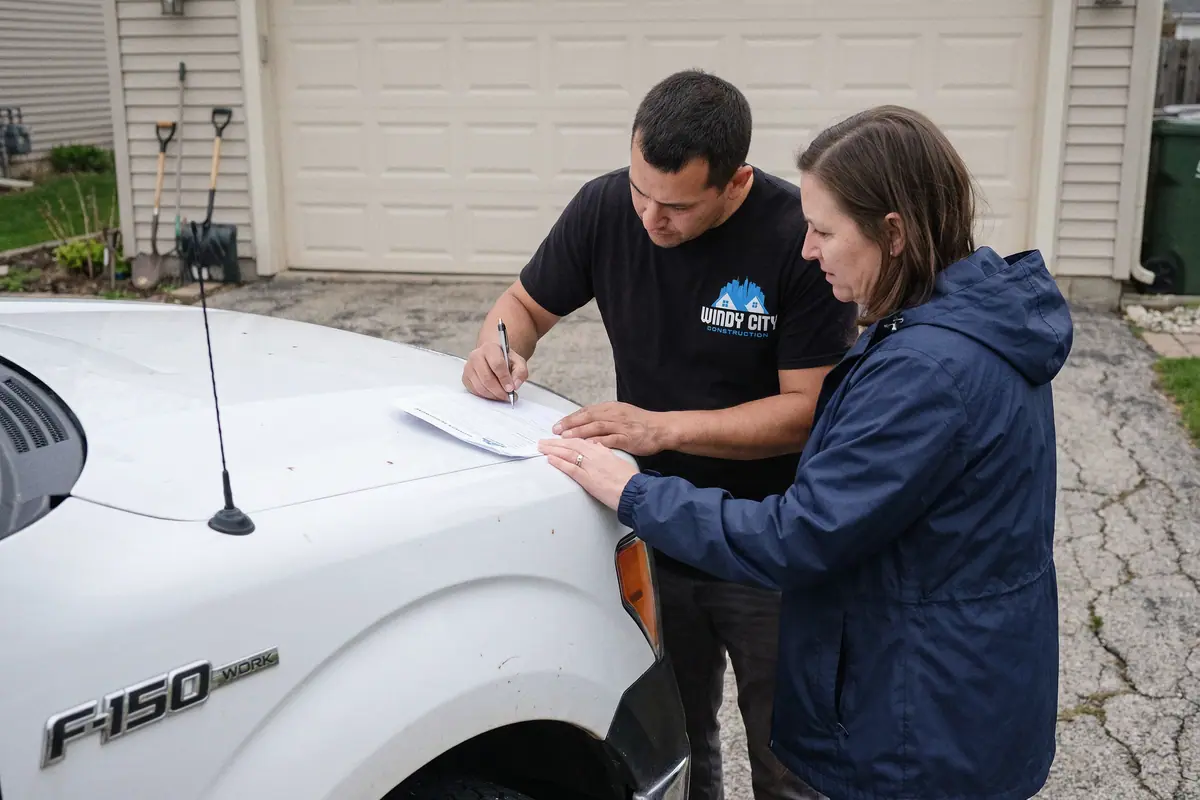 A contractor and a homeowner signing a printed service agreement on the hood of his work truck in her driveway.