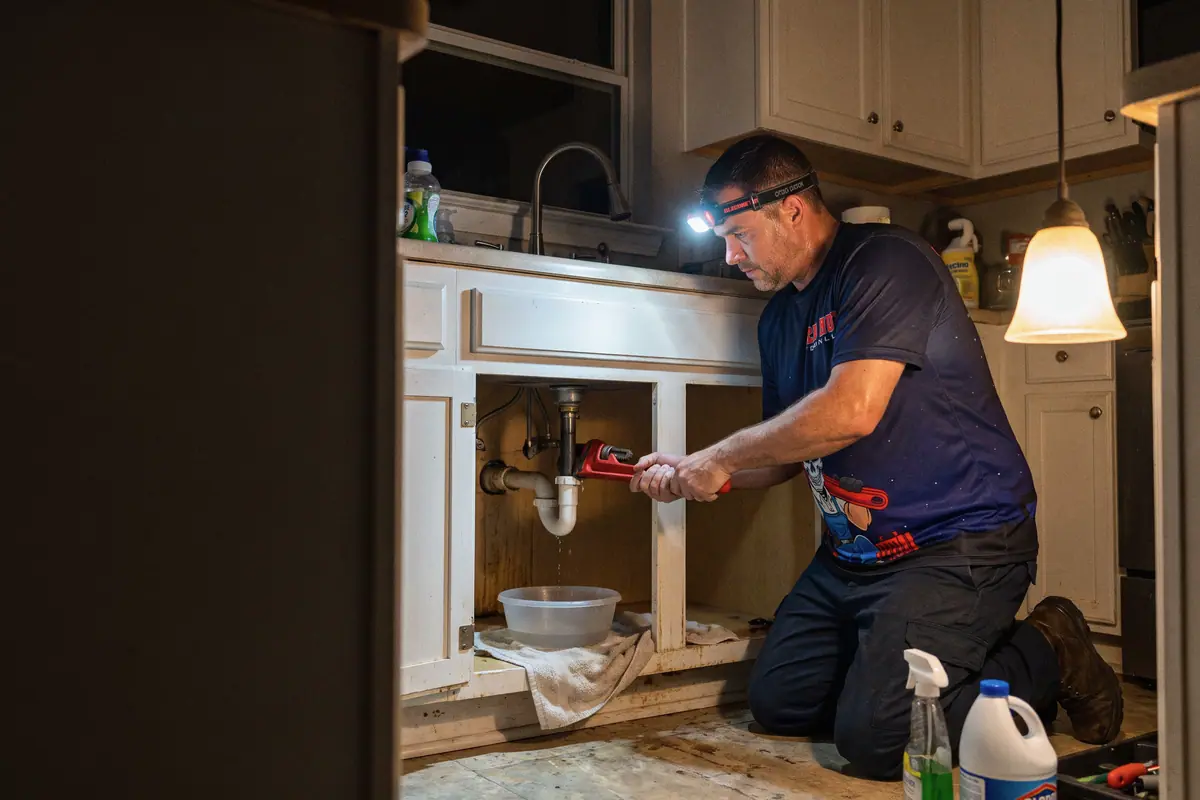 A plumber in a headlamp tightening a leaking pipe under a kitchen sink at night.