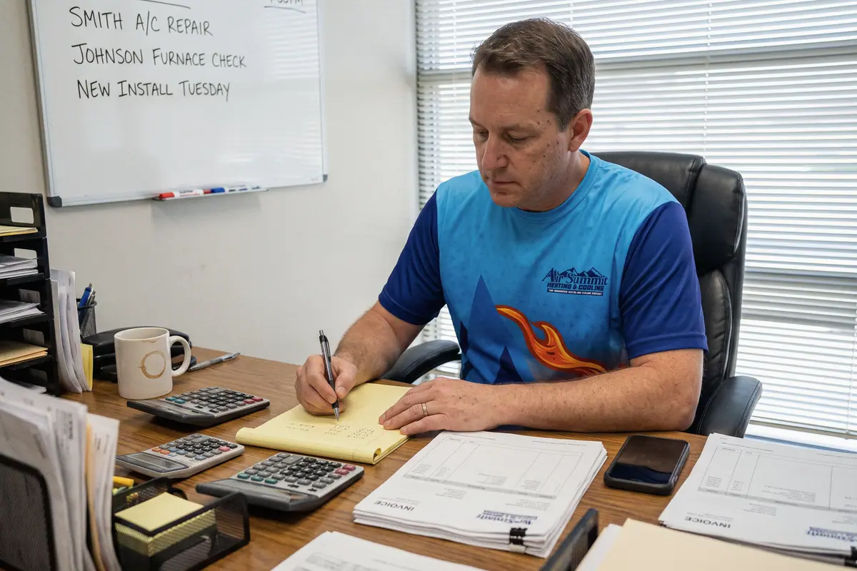 An HVAC owner working out numbers on a legal pad with a calculator at his office desk, the week’s jobs on the whiteboard behind him.