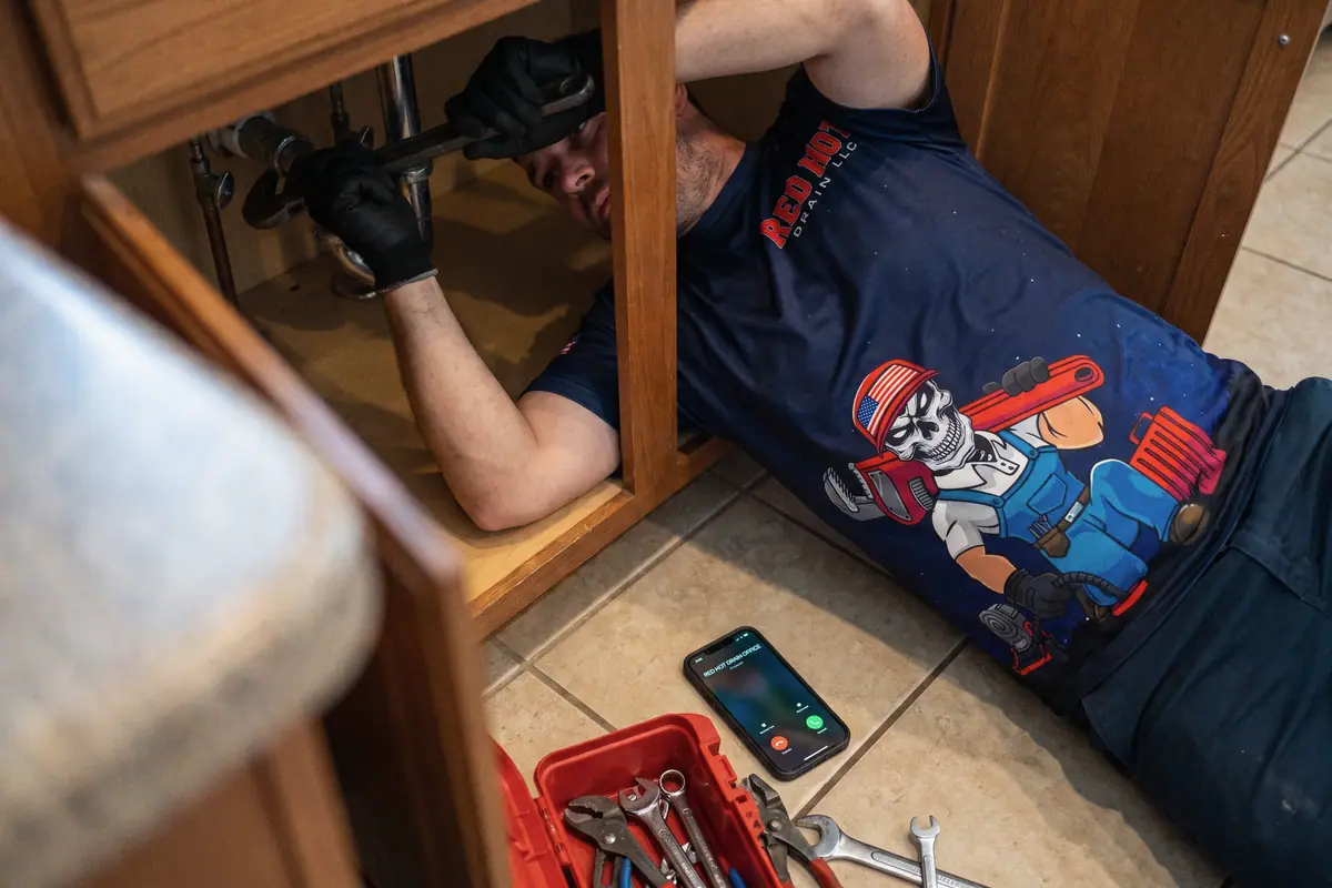 A plumber under a kitchen sink while his phone rings on the floor, out of reach.
