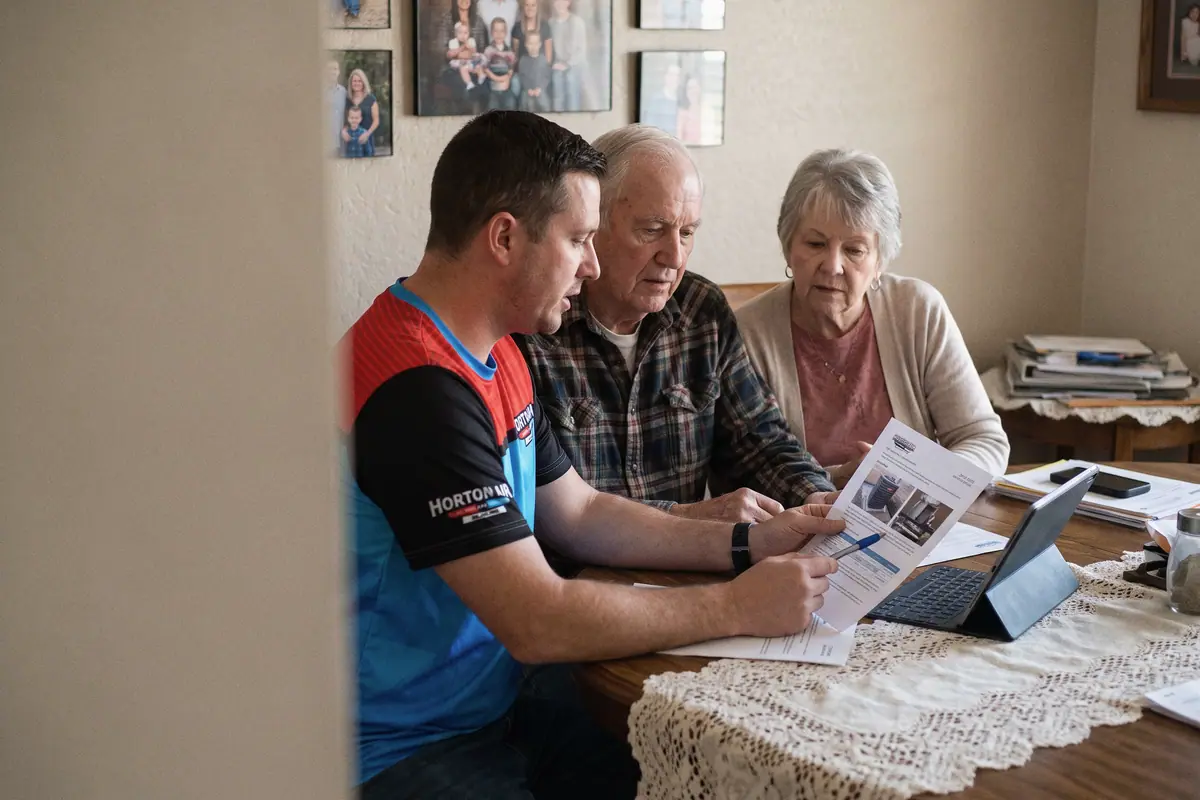 An HVAC technician walking a retired couple through a printed quote at their dining table.